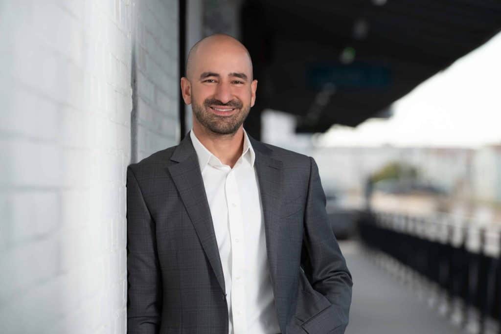 Bright smiling man in business suit leaning against white brick wall at Prosperity CD event.